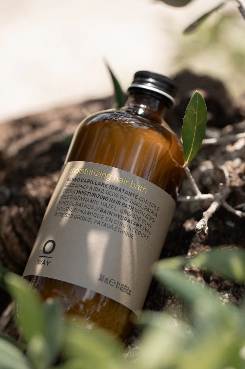 A brown glass bottle of Oway Moisturizing Hair Bath | Hydrating Shampoo for Dry, Lackluster Hair sits on a rock amid green leaves—ideal for restoring moisture and shine to parched hair.