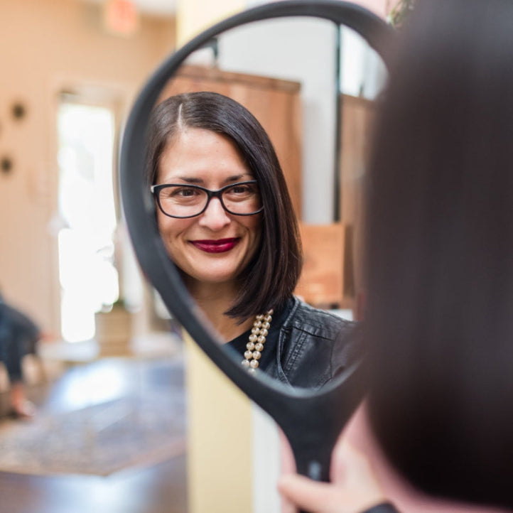 A woman with shoulder-length dark hair and glasses is smiling while looking at her reflection in a handheld mirror inside a room with soft lighting.