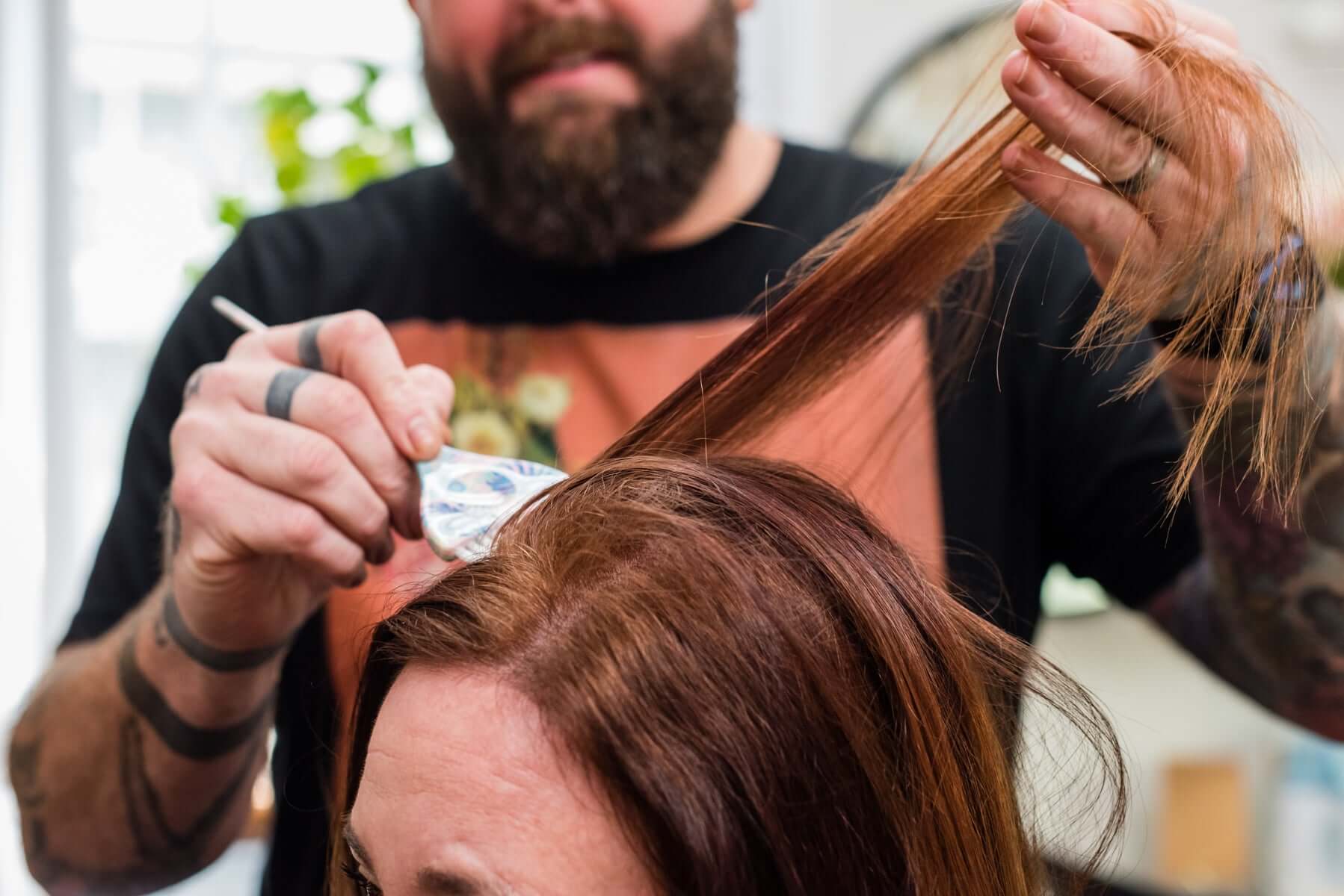A hairdresser with tattoos applies hair dye to a section of a client's brown hair using a brush and foil.