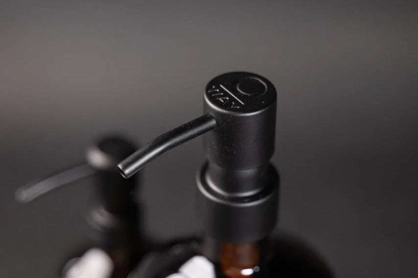 Two Oway Reusable Metal Pump Dispensers from Simply Colour Hair Salon Studio & Online Store sit on a dark surface; one is in front, while the other appears slightly blurred behind. Both are stylish, eco-friendly solutions for daily use.