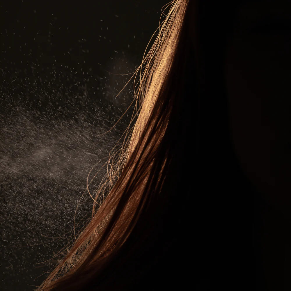 Close-up of light brown hair with fine particles of Oway Molding Mist, a humidity-resistant spray for shape and memory by Oway, suspended in the air against a dark background.