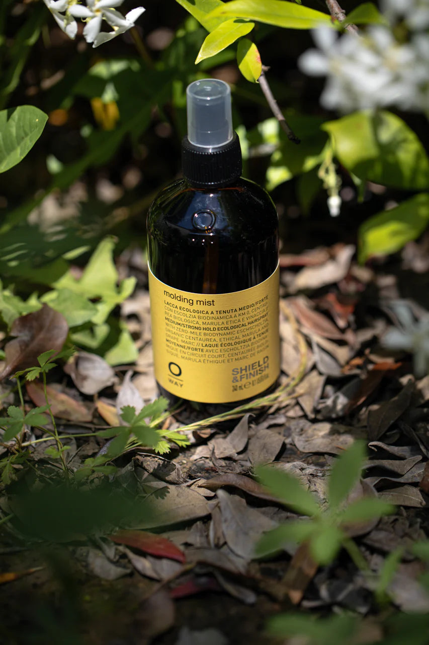 Oway Molding Mist, a humidity-resistant spray with a yellow label, sits among greenery outdoors, providing medium hold and nourishing marula oil for perfect styling.