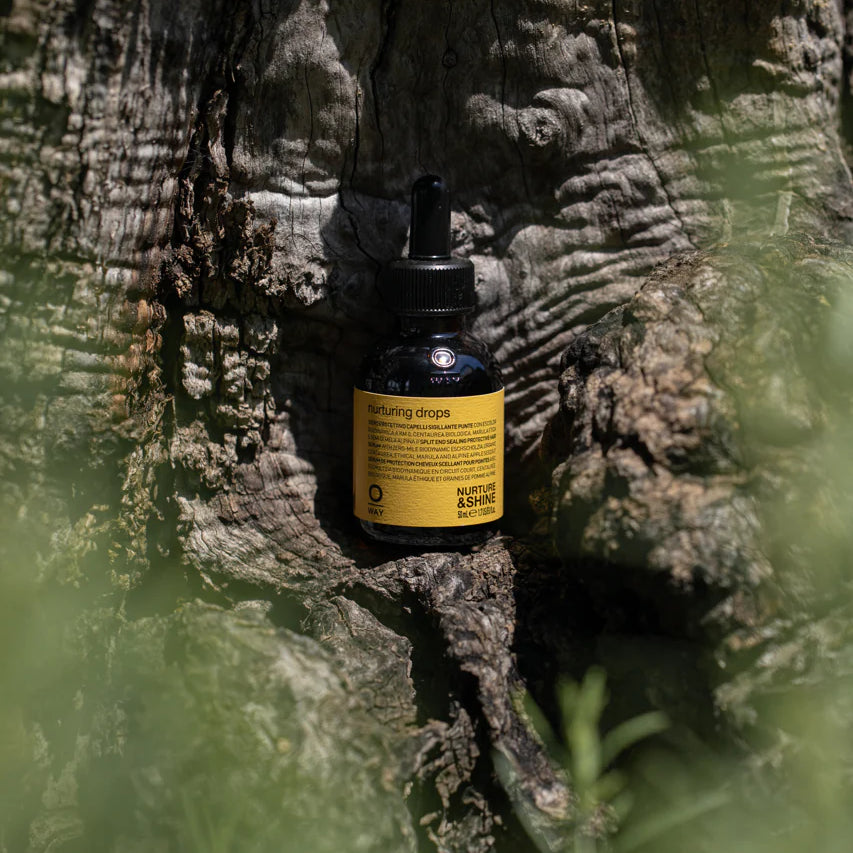 A small dark bottle of Oway Nurturing Drops | Silkening Serum for Split Ends and Heat Protection with a yellow label and dropper cap rests against tree bark, surrounded by green foliage—your go-to for split ends and heat protection.