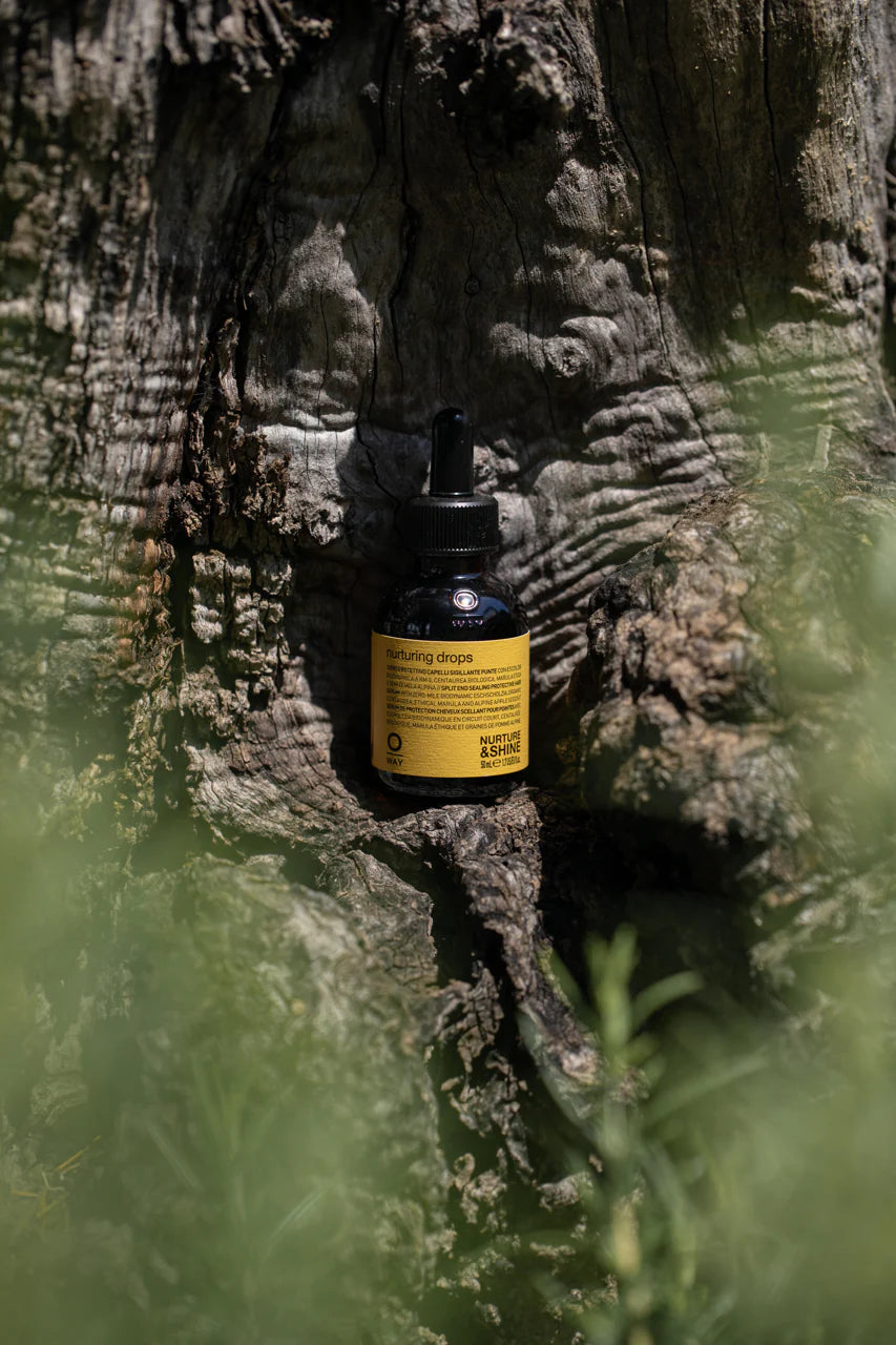 A small dark bottle of Oway Nurturing Drops | Silkening Serum for Split Ends and Heat Protection with a yellow label and dropper cap rests against tree bark, surrounded by green foliage—your go-to for split ends and heat protection.