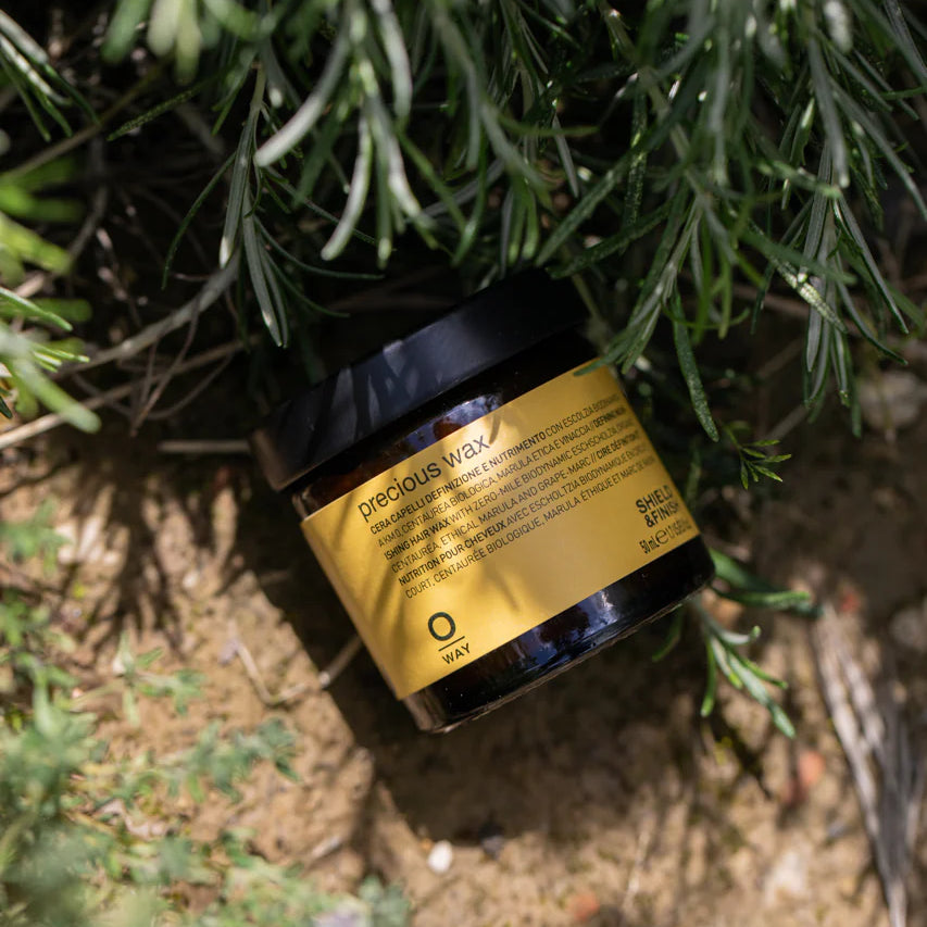 A jar of Oway Precious Wax | Shine Enhancing Styling Wax for Natural Definition with a yellow and black label rests among green plants and soil, highlighting its natural hold and shine-boosting benefits.