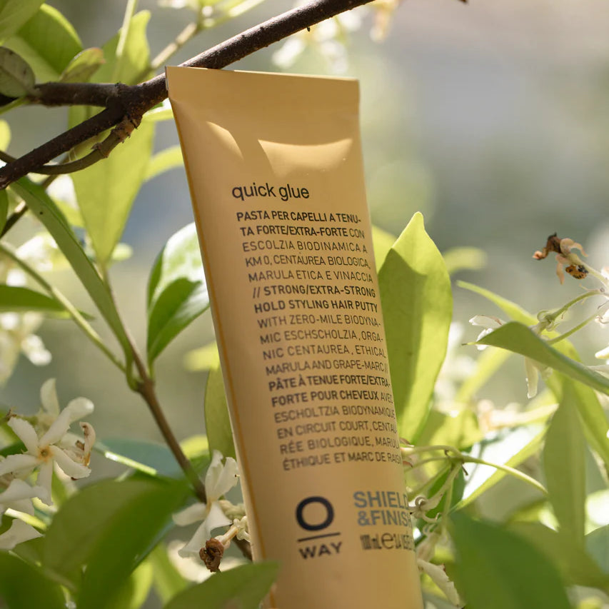 A tube of Oway Quick Glue | Ultra-Hold Styling Glue for Creative Hair Sculpting stands upright among green leaves and white flowers, its label facing outward, showcasing Oway’s glossy-finish formula.