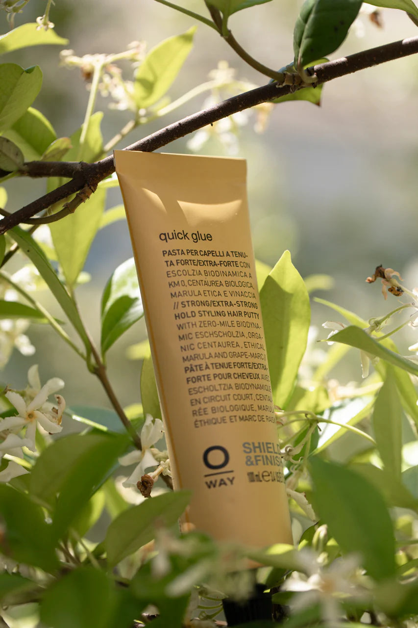 A tube of Oway Quick Glue | Ultra-Hold Styling Glue for Creative Hair Sculpting stands upright among green leaves and white flowers, its label facing outward, showcasing Oway’s glossy-finish formula.