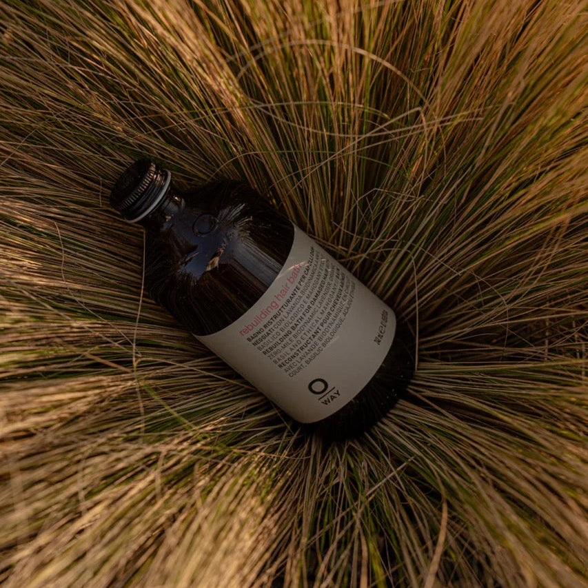 A dark glass bottle with a white label, Oway Rebuilding Hair Bath Strengthening Shampoo for Brittle, Weak Hair, rests on its side amid tall, dry grass.
