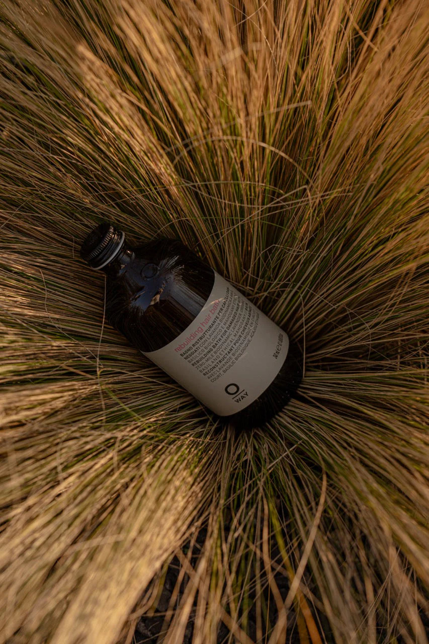A dark glass bottle with a white label, Oway Rebuilding Hair Bath Strengthening Shampoo for Brittle, Weak Hair, rests on its side amid tall, dry grass.
