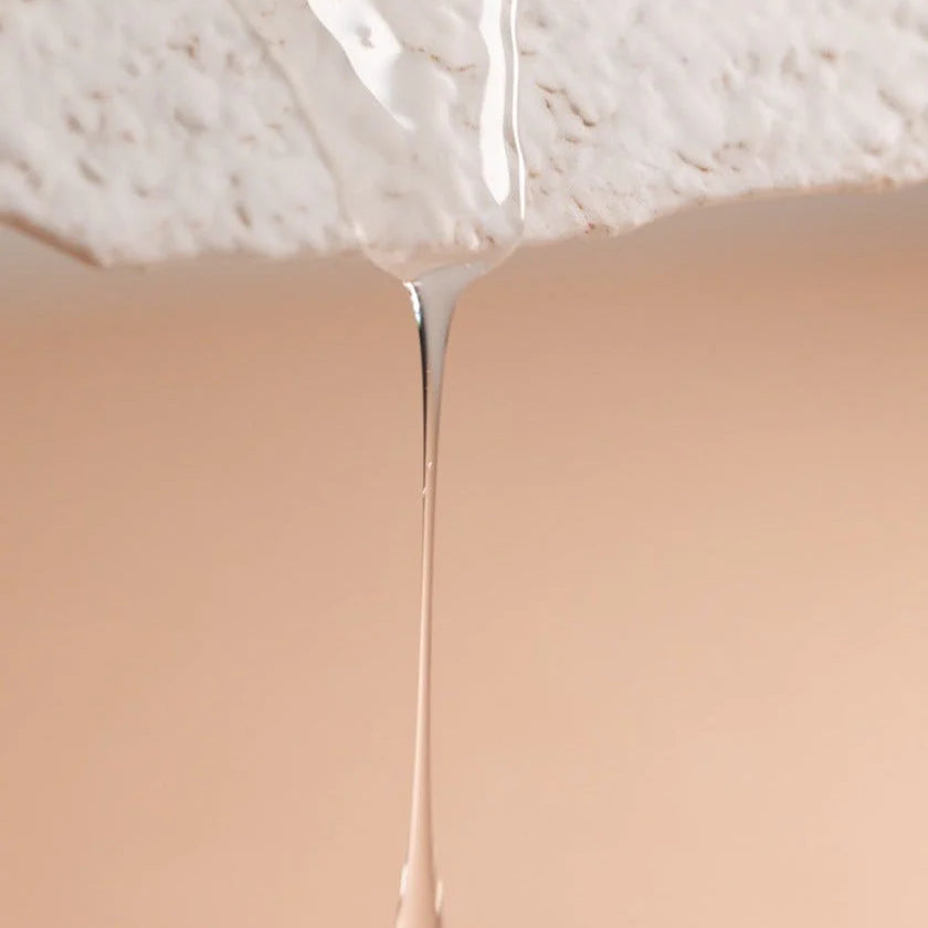 A clear liquid, Oway Rebuilding Hair Bath by Oway—infused with plant-derived keratin—drips from a textured white surface against a beige background, highlighting this strengthening shampoo for brittle, weak hair.