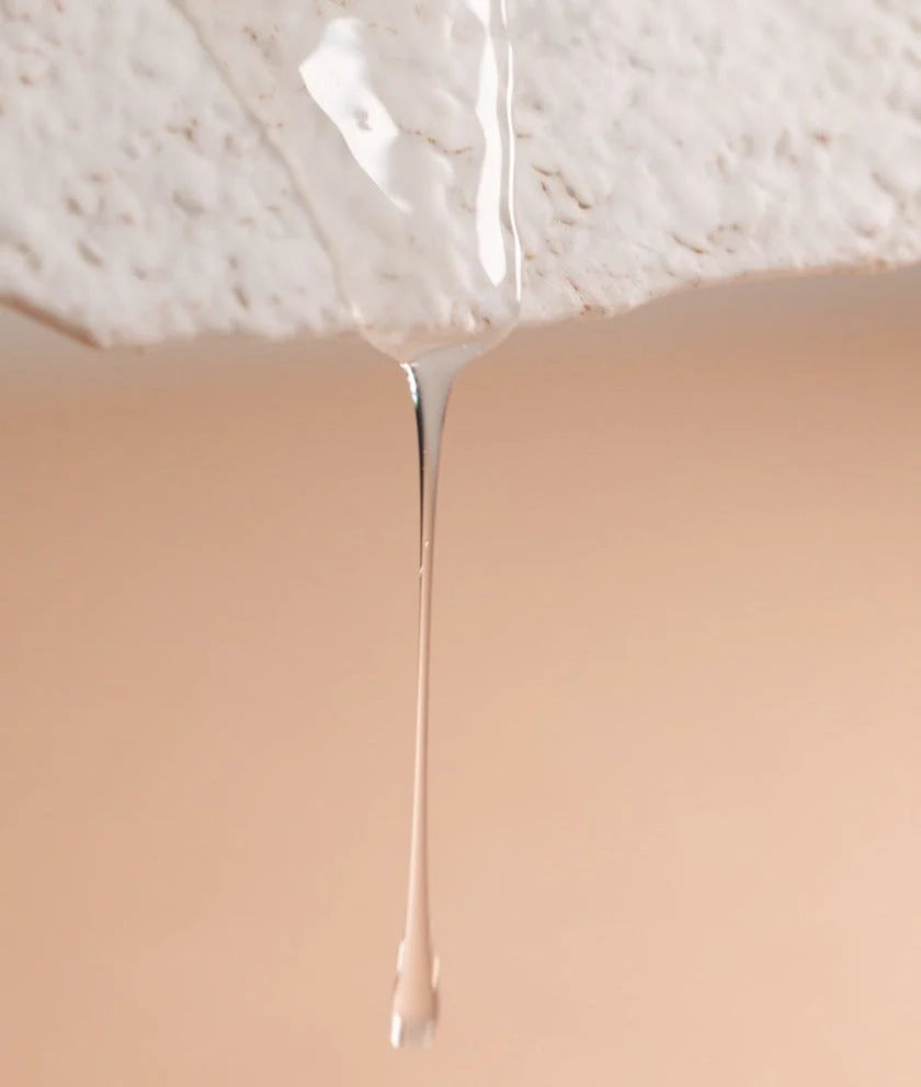 A clear liquid, Oway Rebuilding Hair Bath by Oway—infused with plant-derived keratin—drips from a textured white surface against a beige background, highlighting this strengthening shampoo for brittle, weak hair.