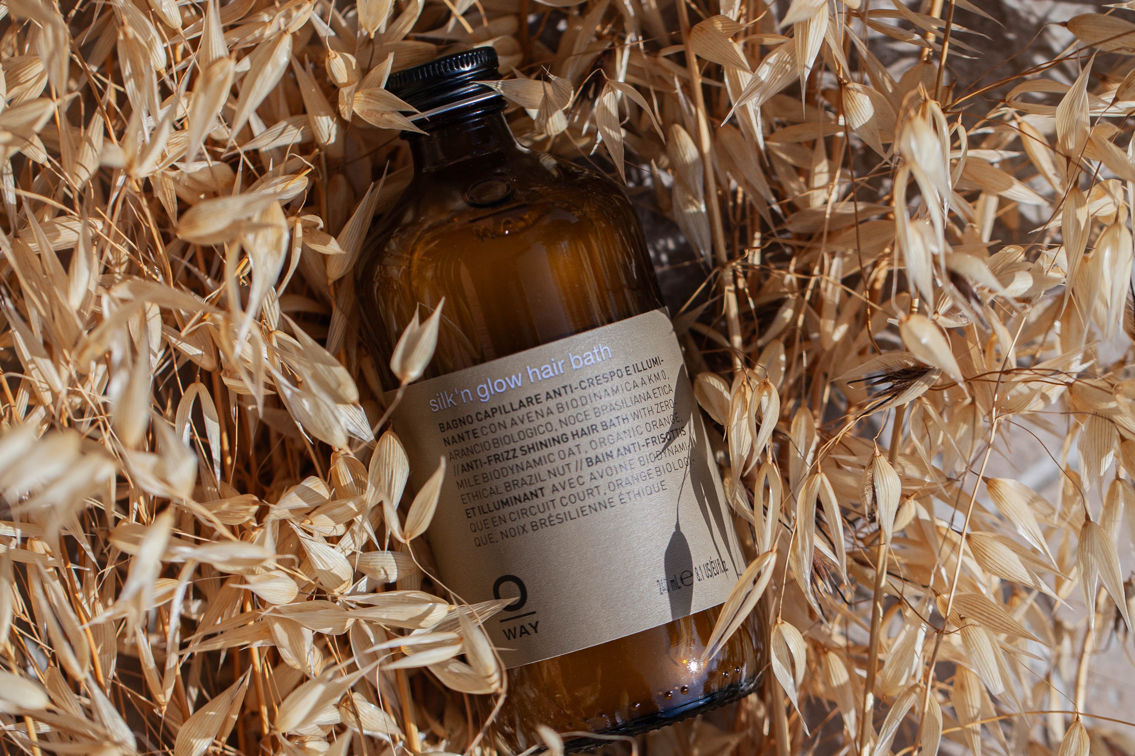 A brown glass bottle labeled Oway Silk’N Glow Hair Bath rests among dried beige grass stalks, sunlight casting shadows—ideal for taming frizz-prone hair.