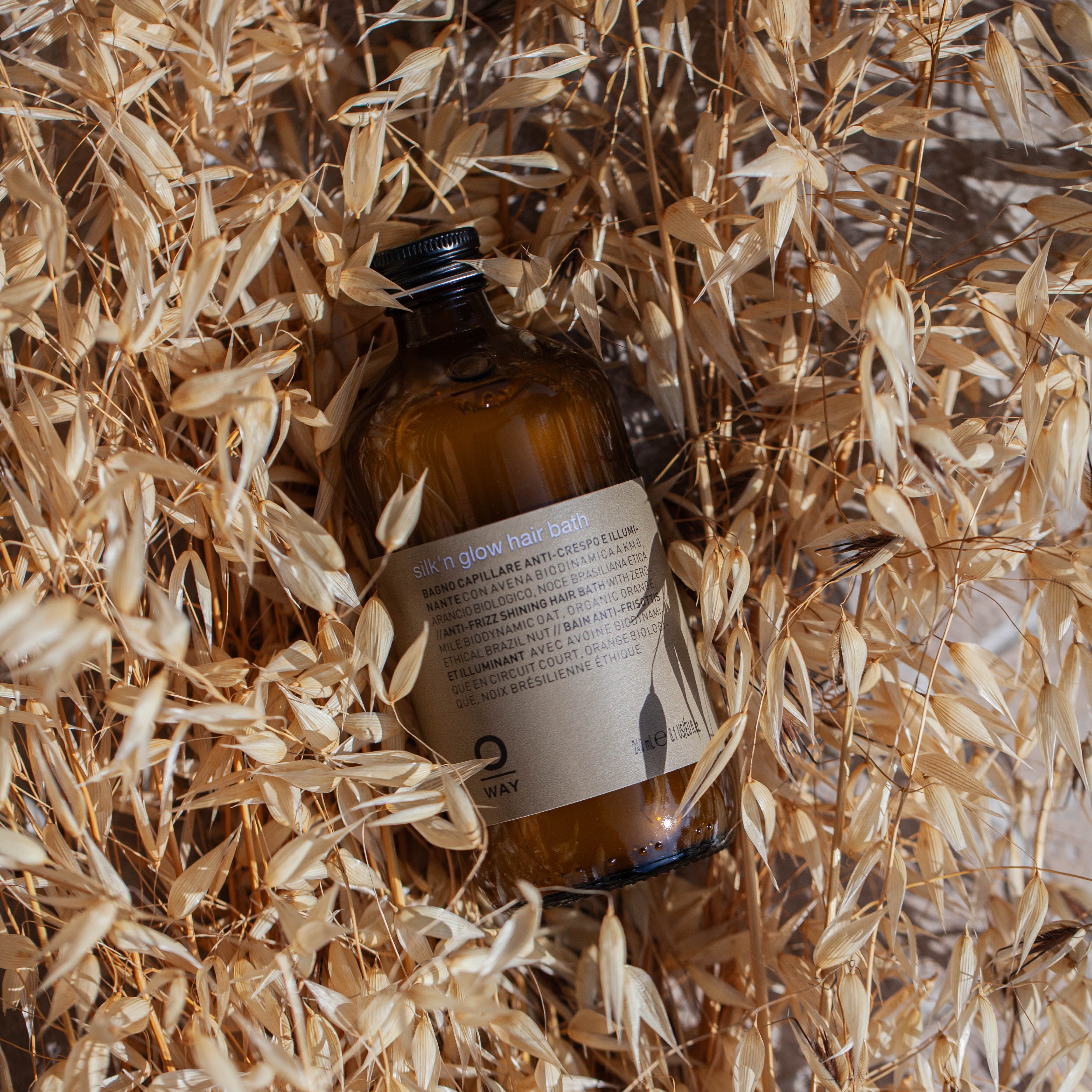 A brown glass bottle labeled Oway Silk’N Glow Hair Bath rests among dried beige grass stalks, sunlight casting shadows—ideal for taming frizz-prone hair.