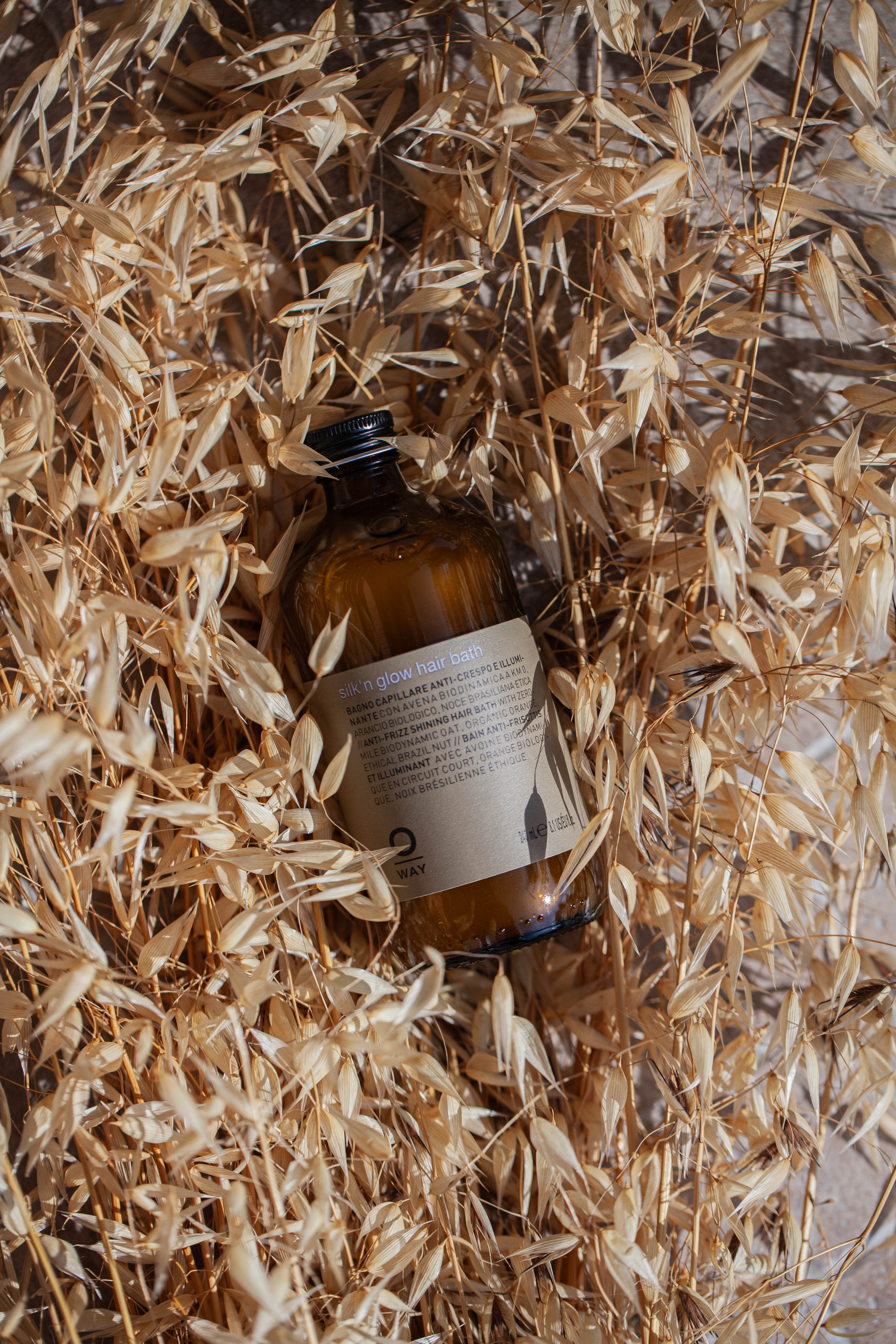 A brown glass bottle labeled Oway Silk’N Glow Hair Bath rests among dried beige grass stalks, sunlight casting shadows—ideal for taming frizz-prone hair.