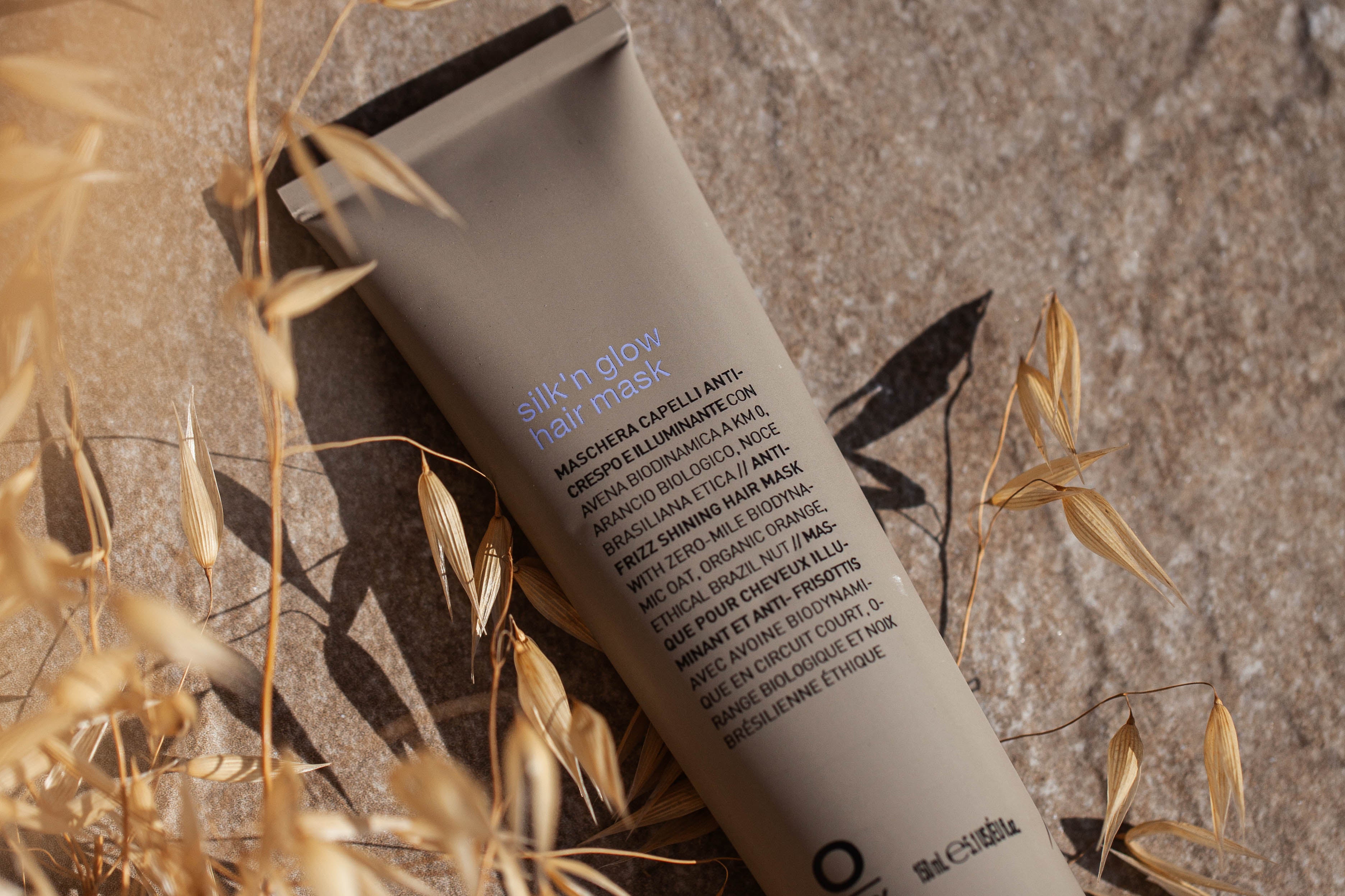 A tube of Oway Silk’N Glow Hair Mask | Nourishing Hair Mask for Dry, Frizzy Hair lies on a stone surface with dried grass and sunlight, ideal for taming dry, frizzy hair with organic Cupuaçu butter.