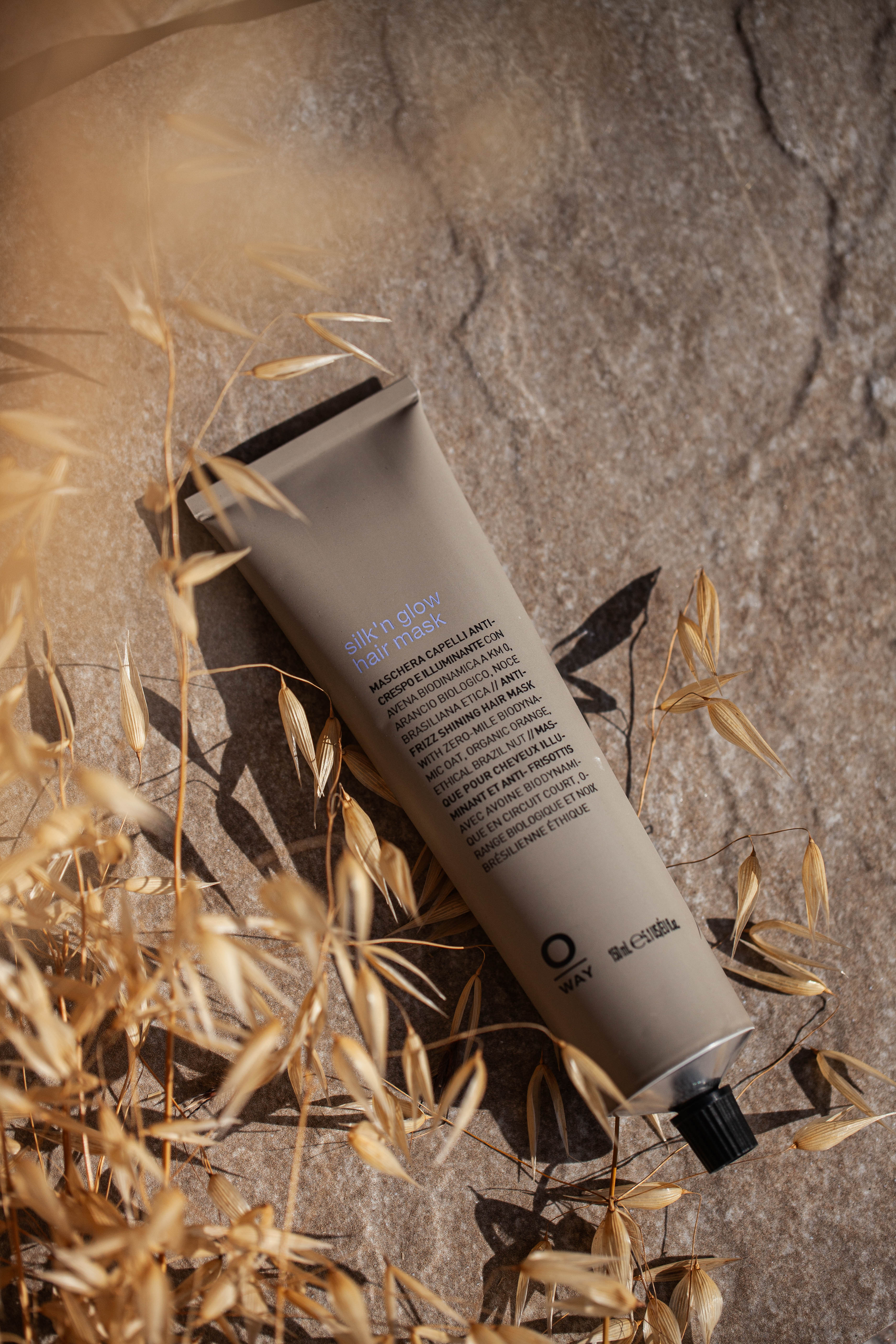 A tube of Oway Silk’N Glow Hair Mask | Nourishing Hair Mask for Dry, Frizzy Hair lies on a stone surface with dried grass and sunlight, ideal for taming dry, frizzy hair with organic Cupuaçu butter.