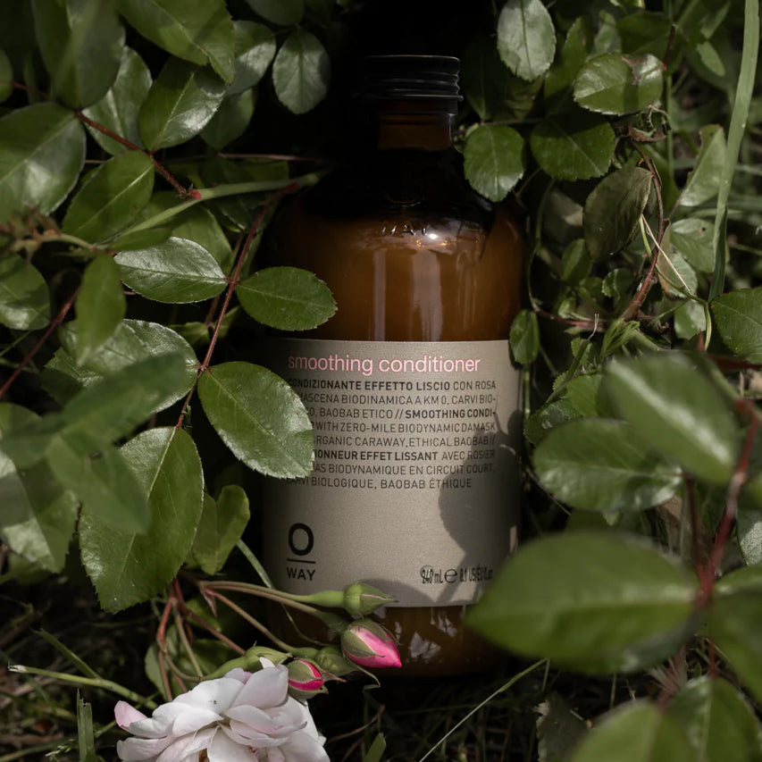 A brown bottle of Oway Smoothing Conditioner | Detangling Conditioner for Soft, Polished Hair sits amid green leaves and grass with a pale pink flower—ideal for taming frizz and enriched with nourishing baobab oil.