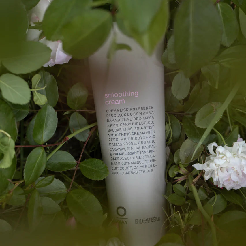 A bottle of Oway Smoothing Cream | Blow Dry Cream for Sleek, Straight Styles is partially hidden among green leaves and white flowers.