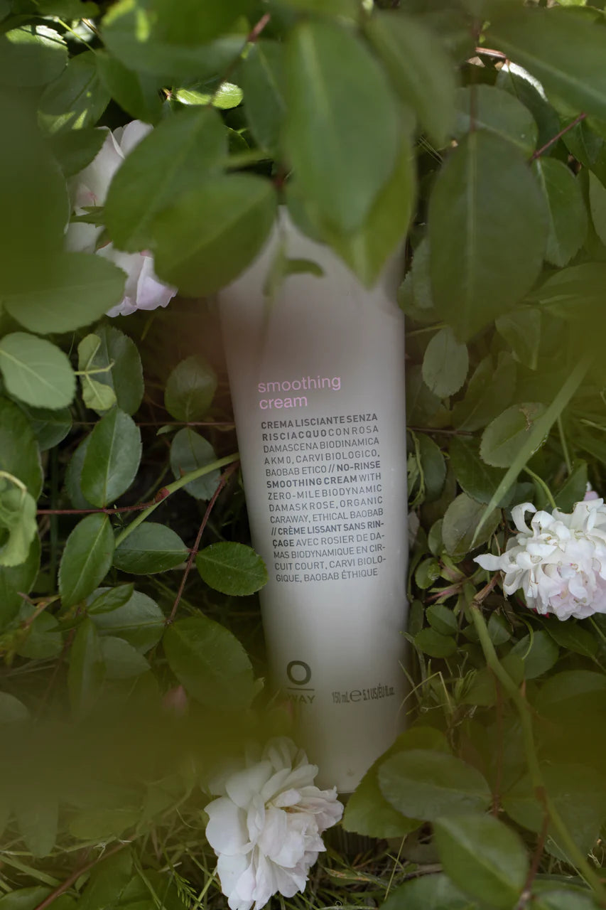A bottle of Oway Smoothing Cream | Blow Dry Cream for Sleek, Straight Styles is partially hidden among green leaves and white flowers.