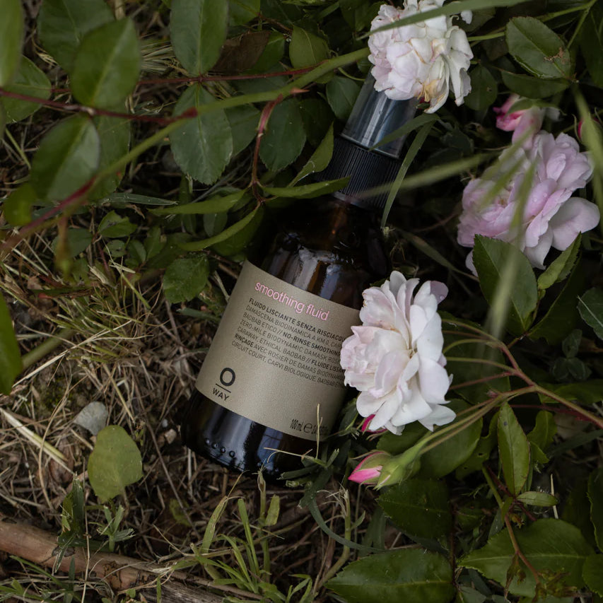 A bottle of Oway Smoothing Fluid | Sleek Styling Serum for Smooth, Frizz-Free Hair rests on grass and soil among green leaves and pale pink roses, promising shiny, sleek hair.