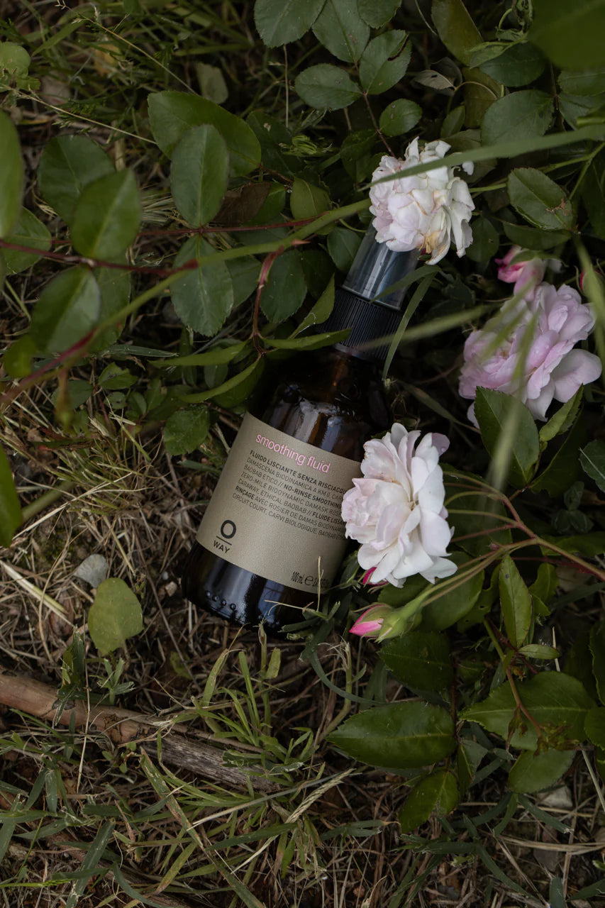 A bottle of Oway Smoothing Fluid | Sleek Styling Serum for Smooth, Frizz-Free Hair rests on grass and soil among green leaves and pale pink roses, promising shiny, sleek hair.