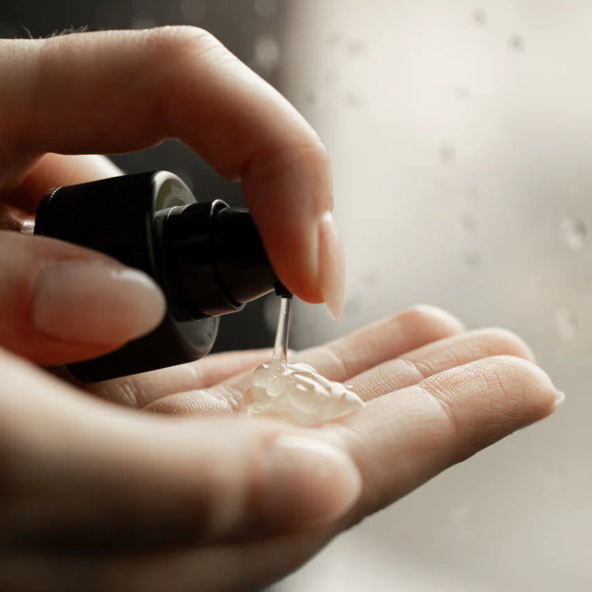 A person dispenses Oway Smoothing Fluid | Sleek Styling Serum for Smooth, Frizz-Free Hair onto their hand, with water droplets on glass behind—perfect for frizz control and enhanced hair shine by Oway.
