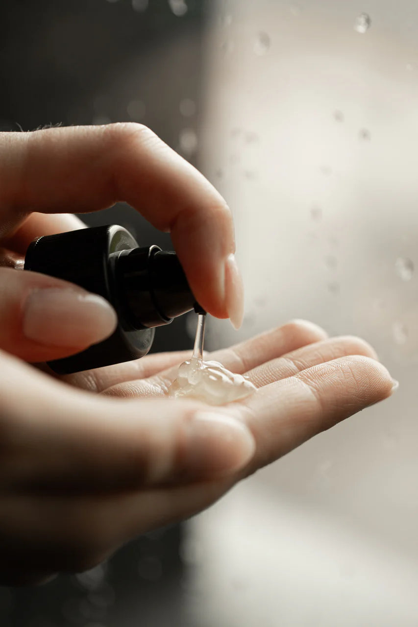 A person dispenses Oway Smoothing Fluid | Sleek Styling Serum for Smooth, Frizz-Free Hair onto their hand, with water droplets on glass behind—perfect for frizz control and enhanced hair shine by Oway.