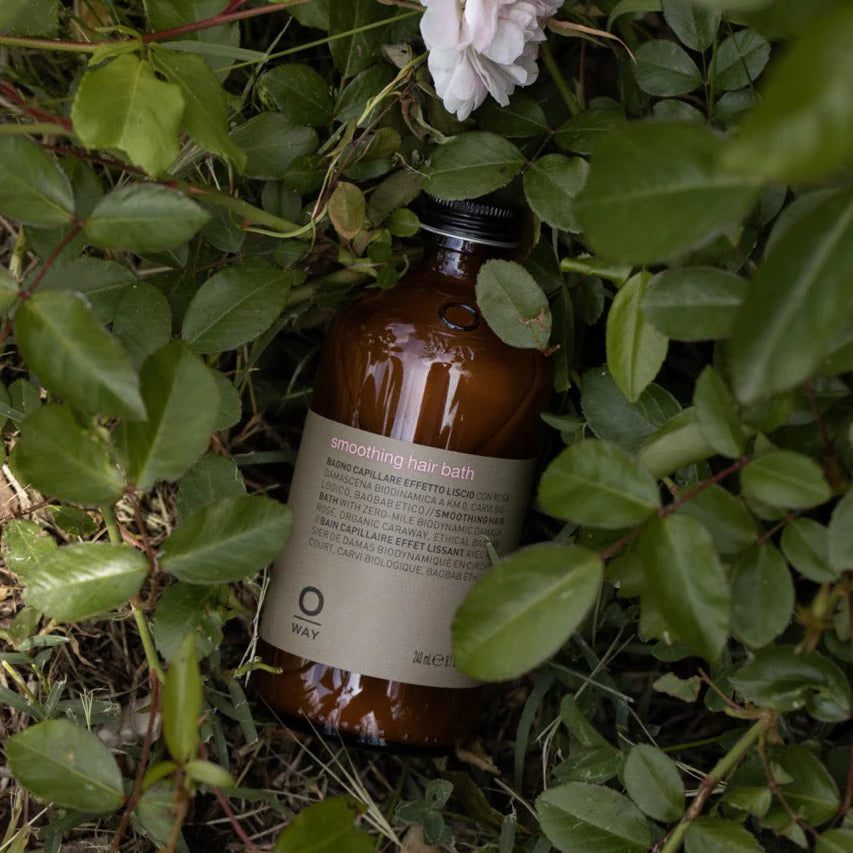 A brown bottle of Oway Smoothing Hair Bath, a frizz-control shampoo for coarse or unruly hair, is nestled among green leaves and an organic rose in a garden setting, suggesting gentle smoothing by Oway.