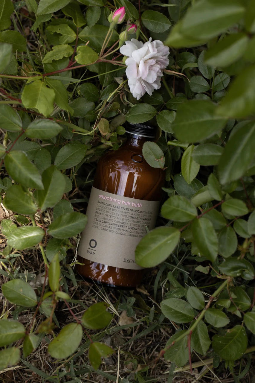 A brown bottle of Oway Smoothing Hair Bath, a frizz-control shampoo for coarse or unruly hair, is nestled among green leaves and an organic rose in a garden setting, suggesting gentle smoothing by Oway.