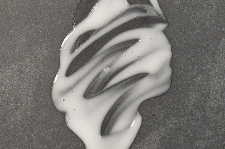 A small amount of Oway Sublime Hair Milk | Lightweight Leave-In Moisture Milk for Softness and Shine by Oway is spilled on a smooth, grey surface, forming an irregular white shape with streaks.