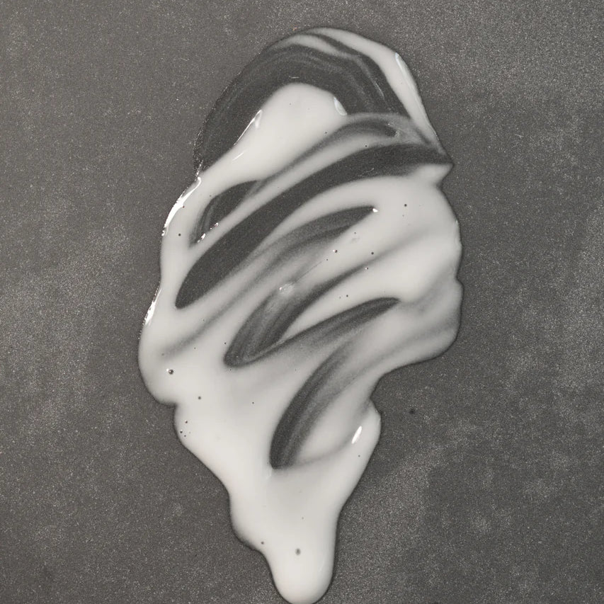 A small amount of Oway Sublime Hair Milk | Lightweight Leave-In Moisture Milk for Softness and Shine by Oway is spilled on a smooth, grey surface, forming an irregular white shape with streaks.