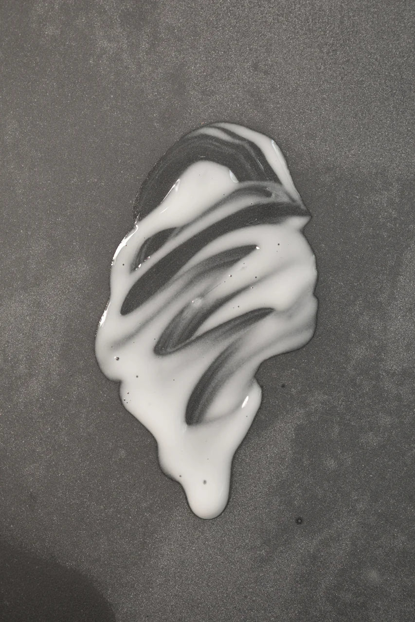 A small amount of Oway Sublime Hair Milk | Lightweight Leave-In Moisture Milk for Softness and Shine by Oway is spilled on a smooth, grey surface, forming an irregular white shape with streaks.