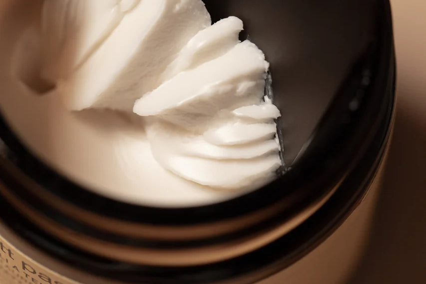 A close-up of Oway Supreme Matte Paste, a strong hold styling paste with a matte finish, as a black spatula scoops some of the product from its brown jar.