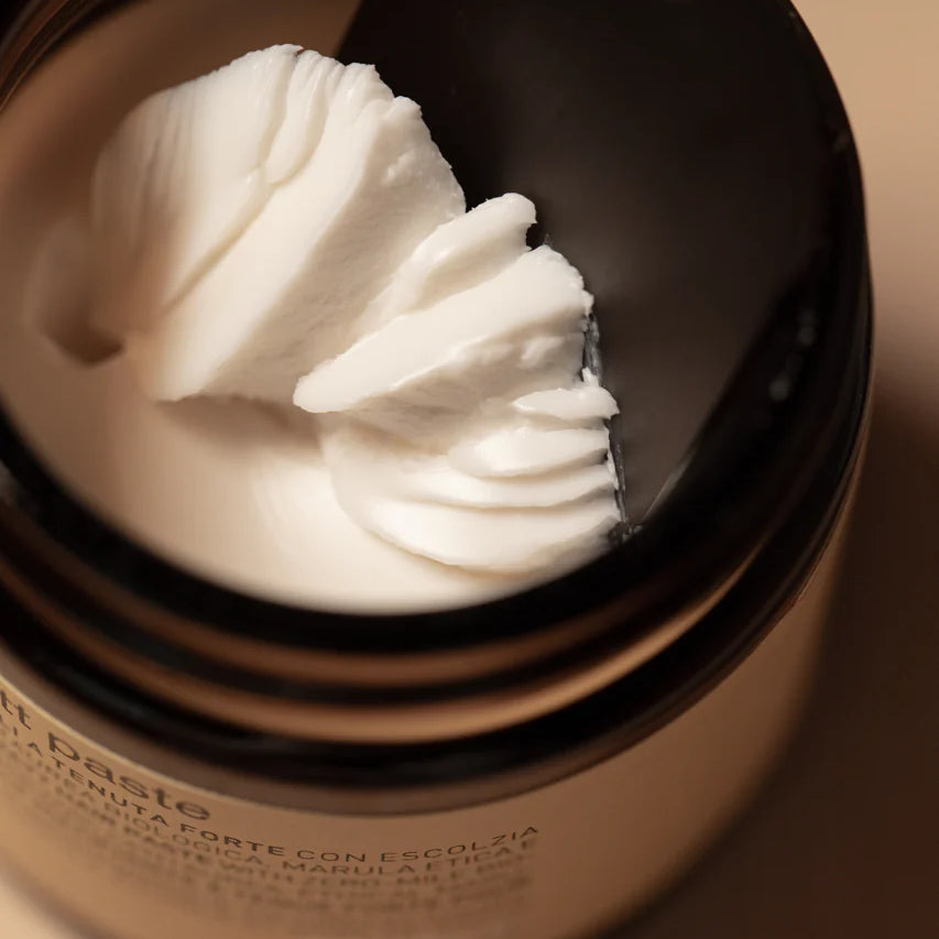 A close-up of Oway Supreme Matte Paste, a strong hold styling paste with a matte finish, as a black spatula scoops some of the product from its brown jar.