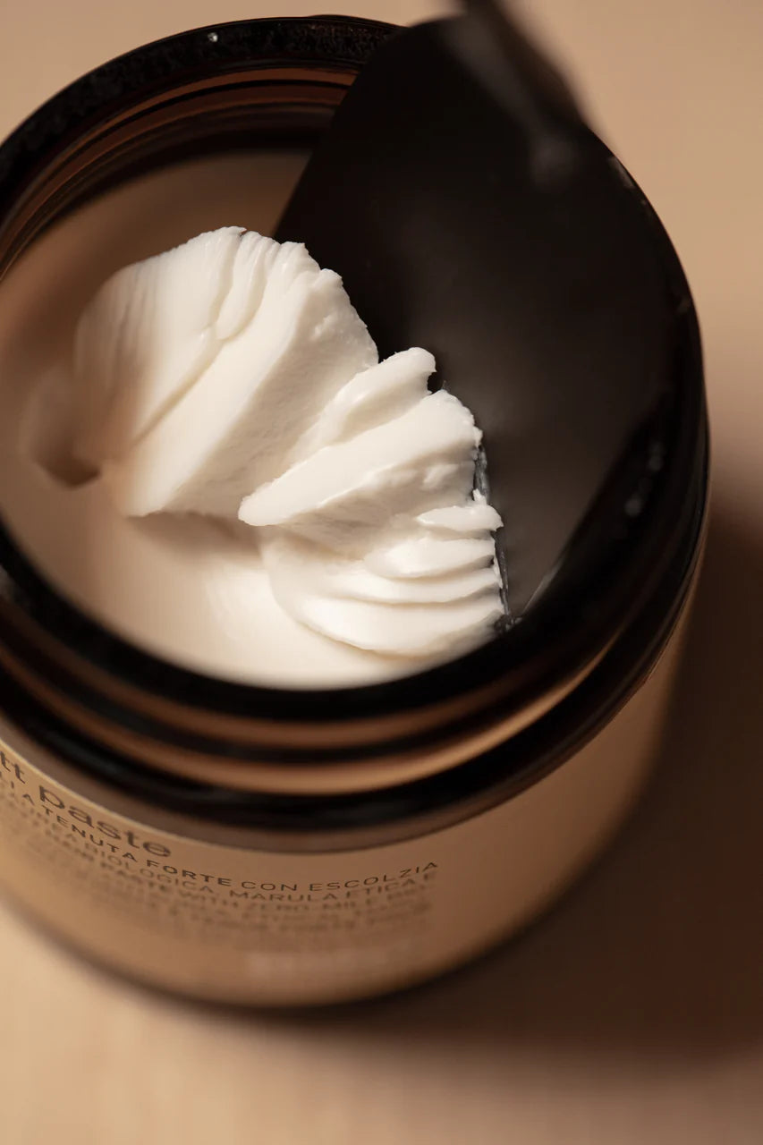 A close-up of Oway Supreme Matte Paste, a strong hold styling paste with a matte finish, as a black spatula scoops some of the product from its brown jar.