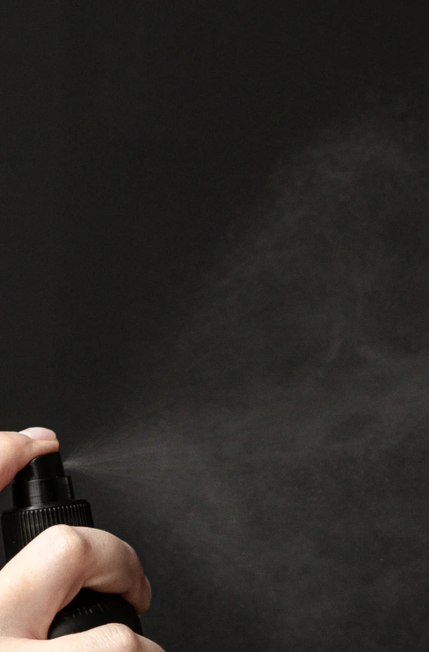 A hand presses the nozzle of a black Oway Flowerfall | Aromatherapeutic Scalp and Mind Balancing Mist bottle, releasing a fine mist against a dark background to evoke holistic scalp and mind care by Oway.