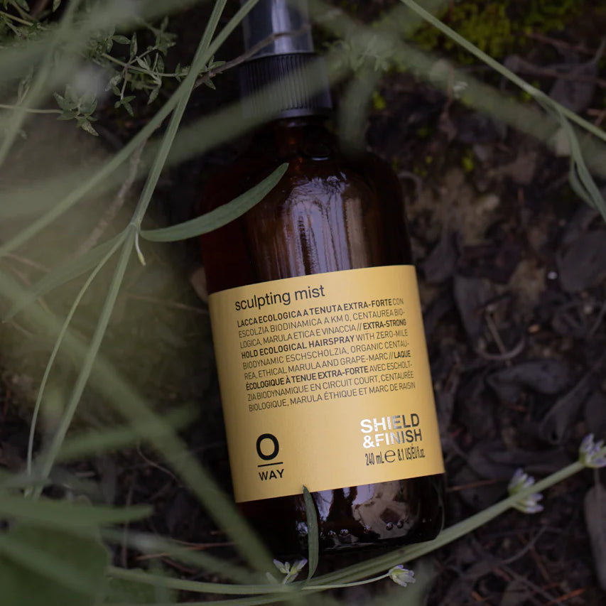The Oway Sculpting Mist, a brown spray bottle, rests among green grass and plants. This long-lasting finishing spray by Oway offers flexible hold and reliable humidity control for your styled hair.