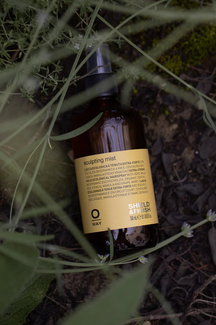 The Oway Sculpting Mist, a brown spray bottle, rests among green grass and plants. This long-lasting finishing spray by Oway offers flexible hold and reliable humidity control for your styled hair.