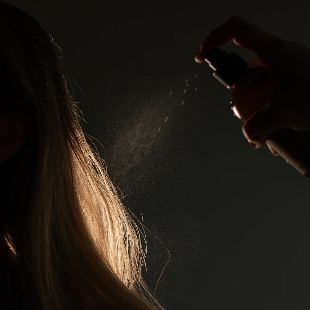 A hand mists Oway Thermo-Detangling Cloud onto long, blonde hair against a dark background, showcasing this Oway heat-protective detangler for all hair types and its essential thermal defense benefits.