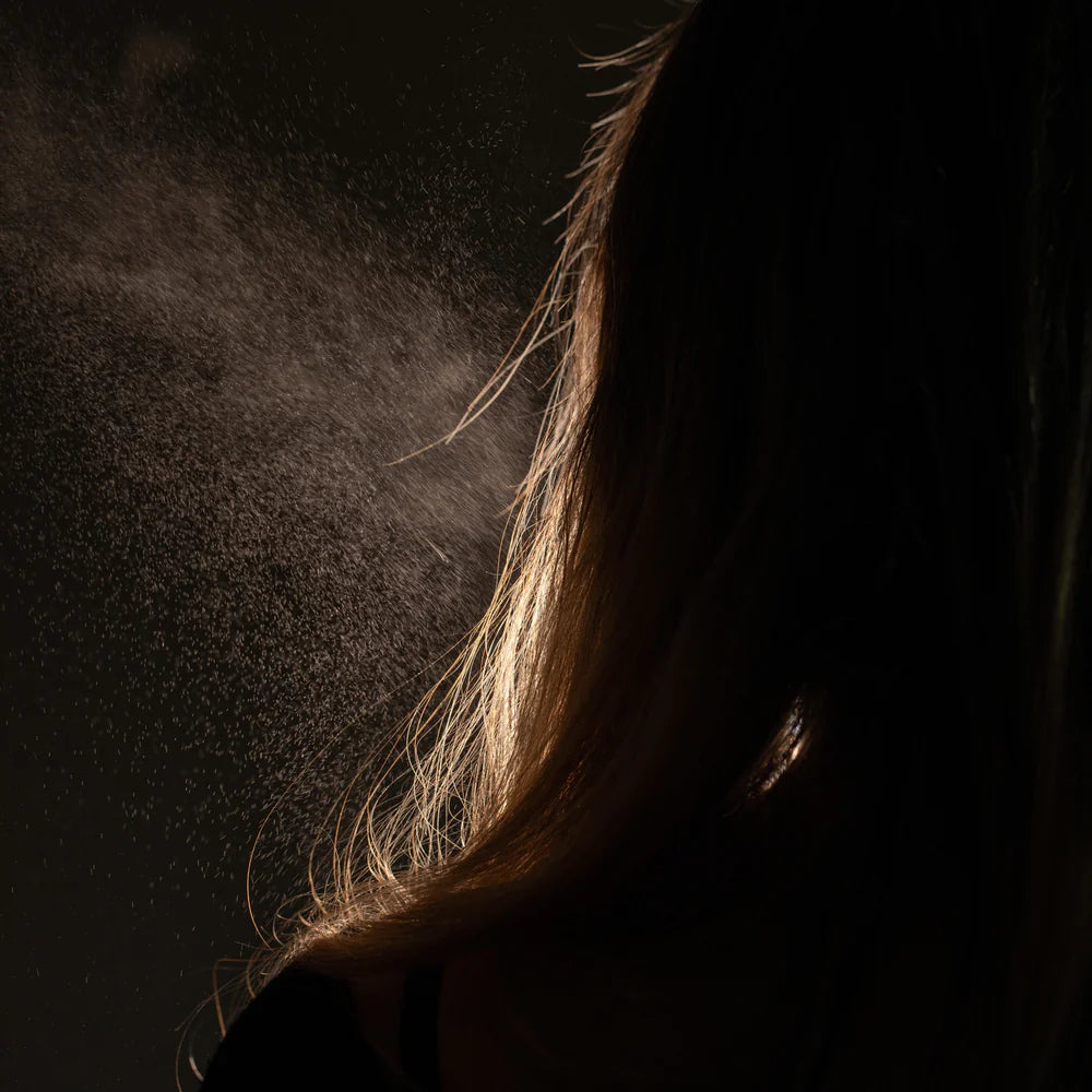 A fine mist of Oway Sea Salt Spray | Beachy Texture Spray for Tousled Waves is applied to long, light brown hair from behind, creating an effortless, tousled look against a dark background.
