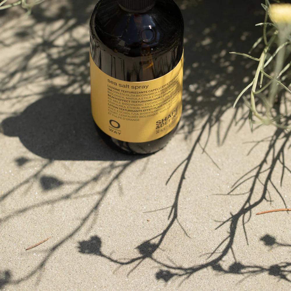 A brown bottle of Oway Sea Salt Spray | Beachy Texture Spray for Tousled Waves sits on sandy ground, plant shadows cast across its label.