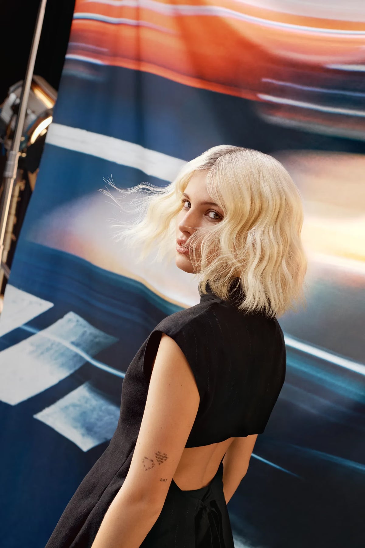 Woman with blonde hair and a black dress standing in front of a colorful abstract painting.