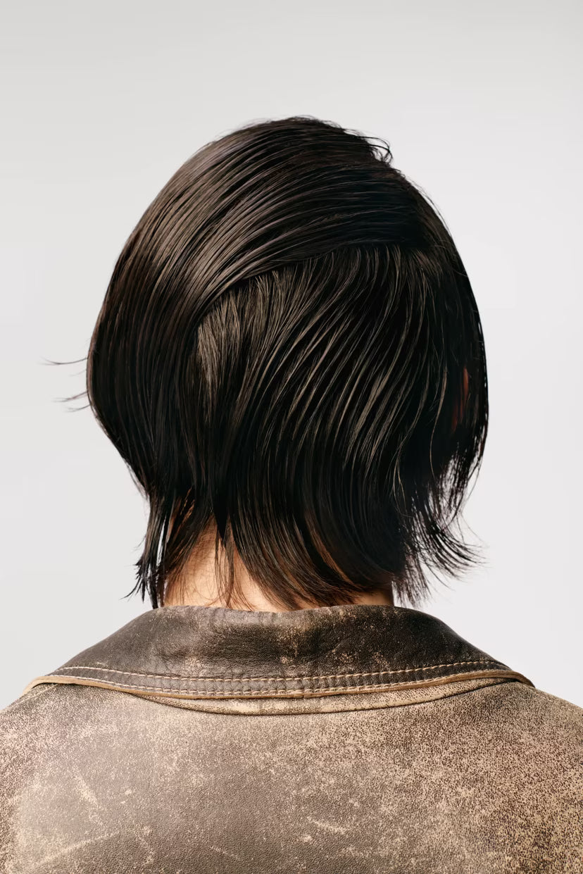 Person with a stylish haircut wearing a textured brown jacket on a light gray background