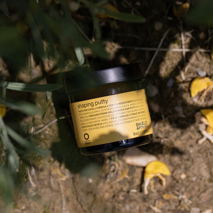 A black Oway Shaping Putty container labeled "Flexible Styling Putty for Sculpted Texture" rests on dirt among green plants and yellow petals, evoking a natural finish inspired by nature.