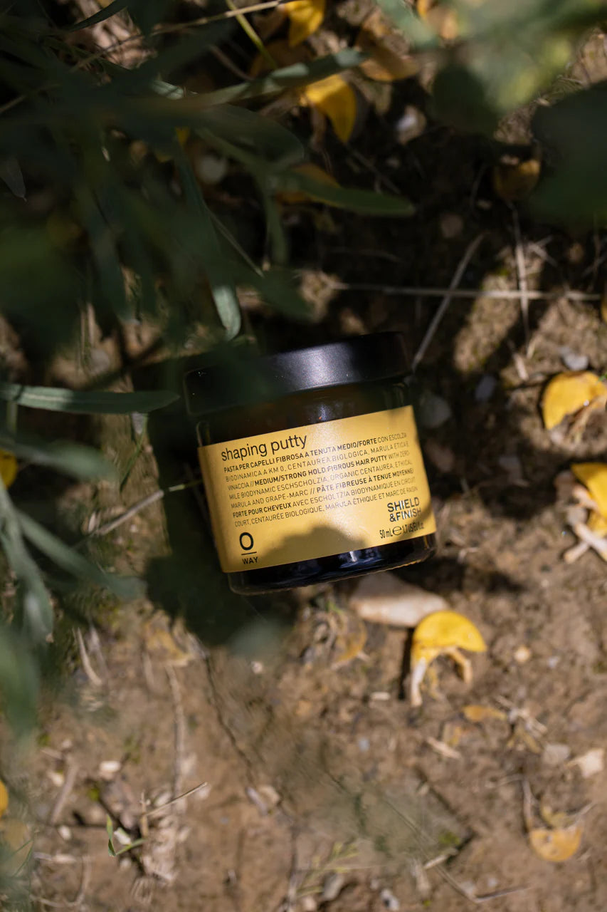 A black Oway Shaping Putty container labeled "Flexible Styling Putty for Sculpted Texture" rests on dirt among green plants and yellow petals, evoking a natural finish inspired by nature.