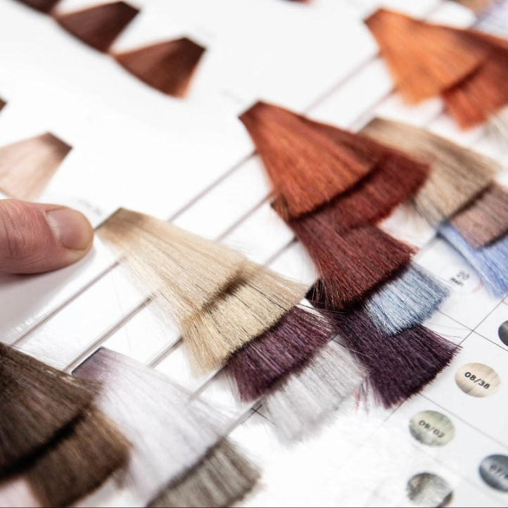 A close-up of a person's finger pointing at a sample of light blonde hair on a hair color swatch chart, with various other hair color samples visible on the page.