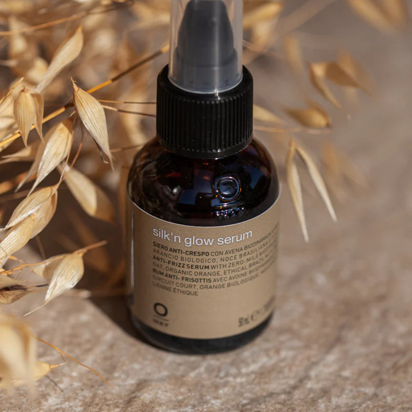 A bottle of Oway Silk’N Glow Serum | Anti-Frizz Shine Serum for Dry, Frizzy Hair with a pump dispenser rests on a textured surface beside dried oat stalks, showcasing its nourishing Brazil nut oil and biodynamic oat infusion.