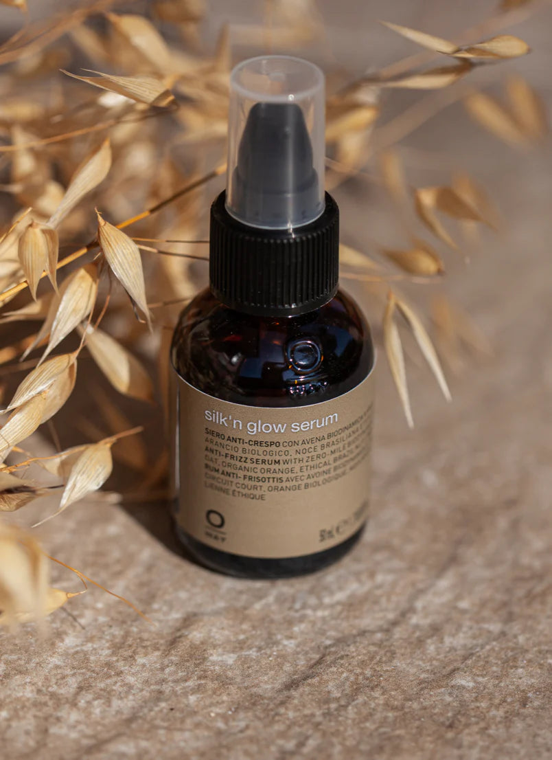 A bottle of Oway Silk’N Glow Serum | Anti-Frizz Shine Serum for Dry, Frizzy Hair with a pump dispenser rests on a textured surface beside dried oat stalks, showcasing its nourishing Brazil nut oil and biodynamic oat infusion.