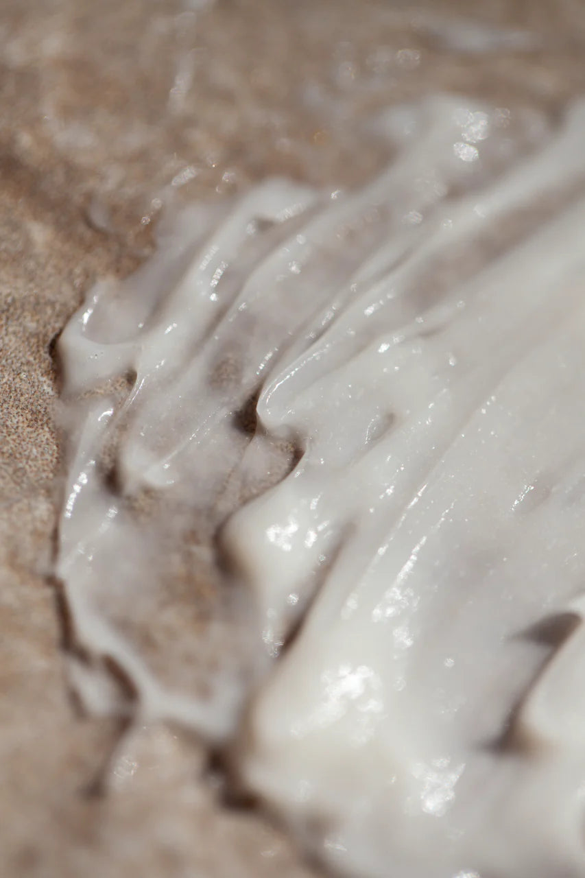 A close-up of Oway Smoothing Cream | Blow Dry Cream for Sleek, Straight Styles by Oway, featuring its creamy white texture with biodynamic caraway spread smoothly on a textured brown surface.