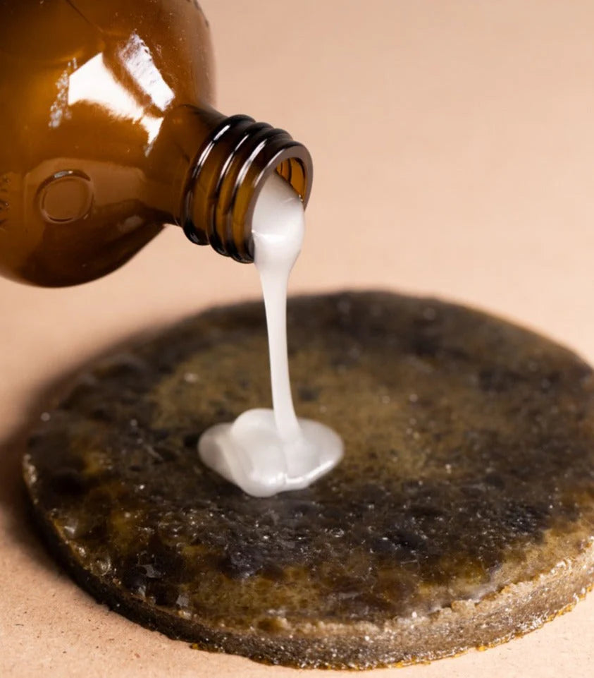 A brown Oway bottle pours Smoothing Hair Bath—an anti-frizz shampoo for coarse or unruly hair—onto a dark textured surface with a tan backdrop, perfect for smoothing and taming frizzy hair.